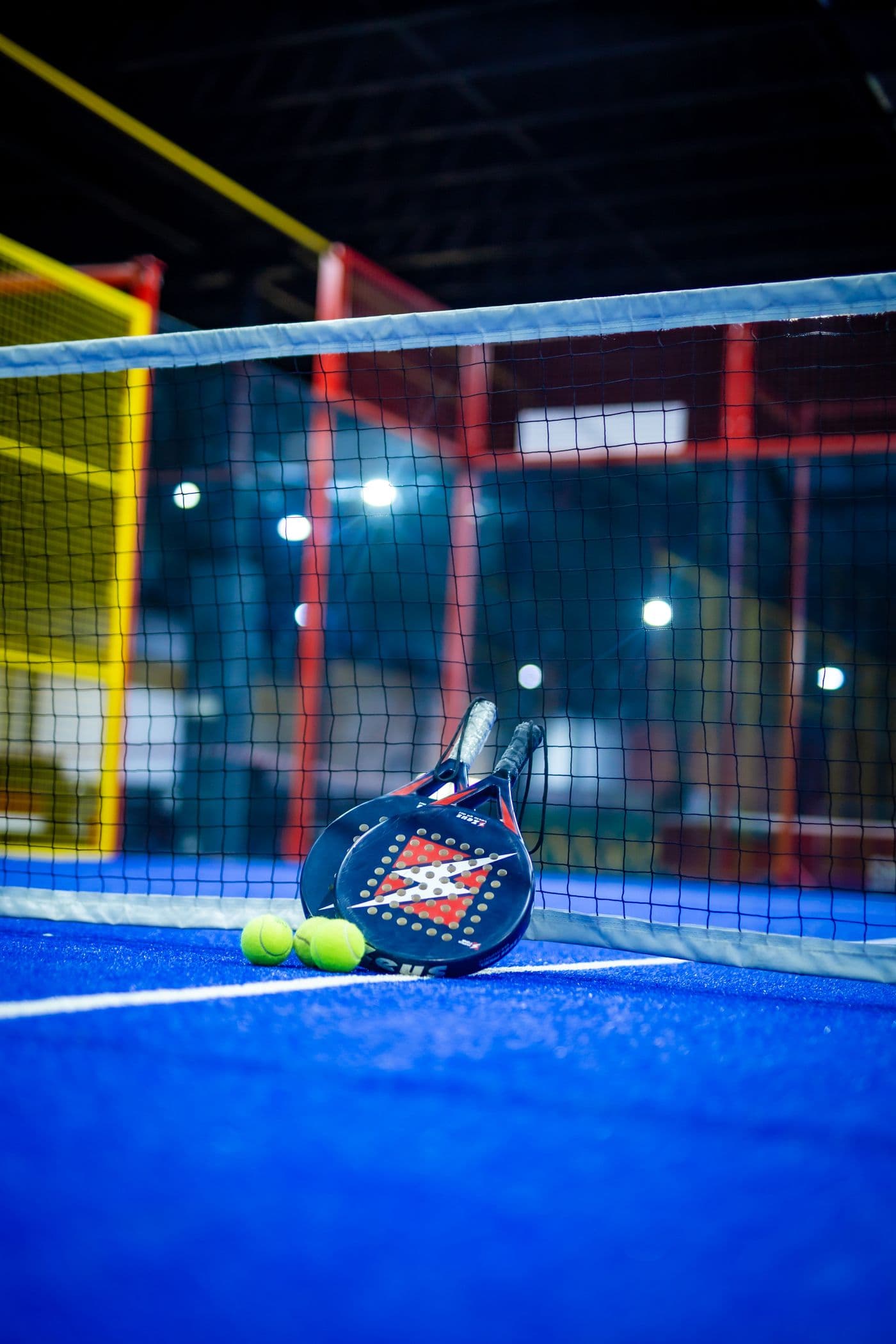 Two Padel rackets and balls leaning against the net on a blue court under floodlights